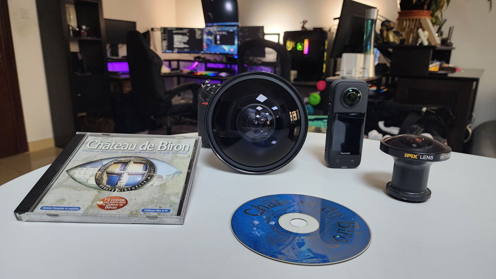 The 1999 Château de Biron CD-ROM and box, a Nikkor fisheye on a Nikon, an iPIX lens adapter and an Insta360 camera on a white table.